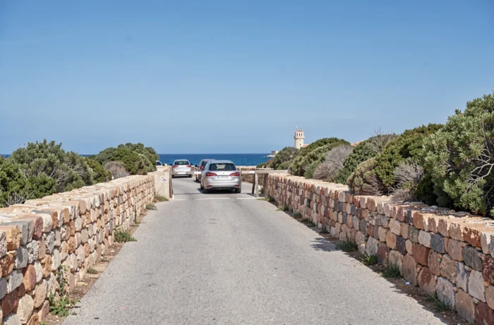 Route d'accès au parking de la plage La Pelosa à Stintino avec voitures en attente et tour historique en arrière-plan